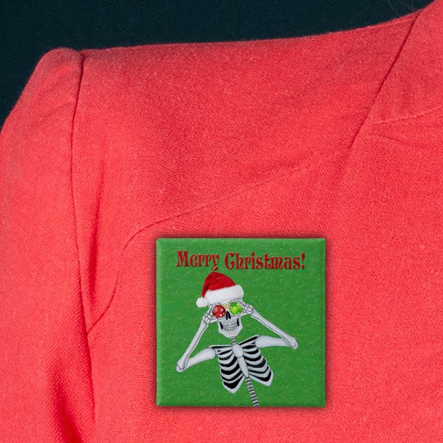 Frohe Weihnachten Glück Skeleton Weihnachtsmannmüt Button (Happy Christmas skeleton holding ornaments in Santa Hat on green with gold flecks pin back button.)