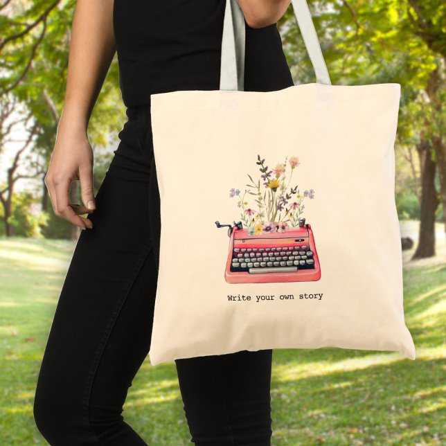 Schreiben Sie Ihre eigene Geschichte, Schriftstell Tragetasche (A woman holding a tote bag with a typewriter graphic and wildflowers & text: "write your own story")