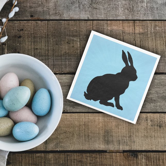 Schwarzer Bunny Silhouette Form Tall Ohren auf bla Serviette (Sitting black silhouette bunny rabbit on blue paper napkin for Easter or spring time.)