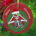 Skelett in Pine Christmas Wreath Red Hat Funny Keramik Ornament<br><div class="desc">Dieses hängende Keramik-Ornament hat ein lustiges Skelett mit Schneeflocken und einer roten Weihnachtsmannmütze in einem grünen Button auf tiefrotem Hintergrund. Passen Sie Ihren Text auf der Rückseite mit dem und das Frohe Weihnachten auf der Vorderseite mit den Vorlagen an.</div>