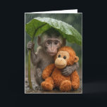 Thinking of You Macaque Monkey with Orangutan Karte<br><div class="desc">A young macaque monkey uses a large green leaf as a makeshift umbrella while holding onto a stuffed orange orangutan toy in the gentle rain of a lush forest.
Inside text can be changed.</div>